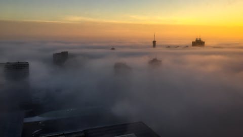 Het is lastig te voorspellen of de zon mist wel of niet oplost. Uit een wolkendek steekt hoogbouw. Op de achtergrond is de opkomende zon te zien.