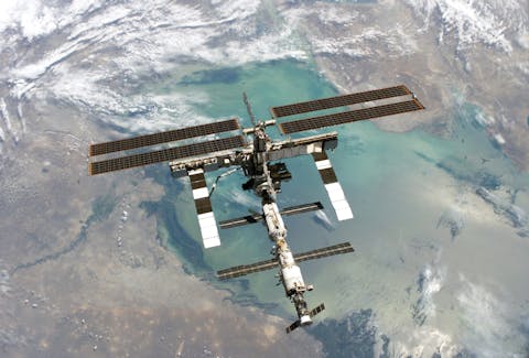 Het internationale ruimtestation ISS in aanbouw in 2005. Een ruimtestation met zonnepanelen vliegt boven de aarde. Land, zee en wolken zijn te zien.