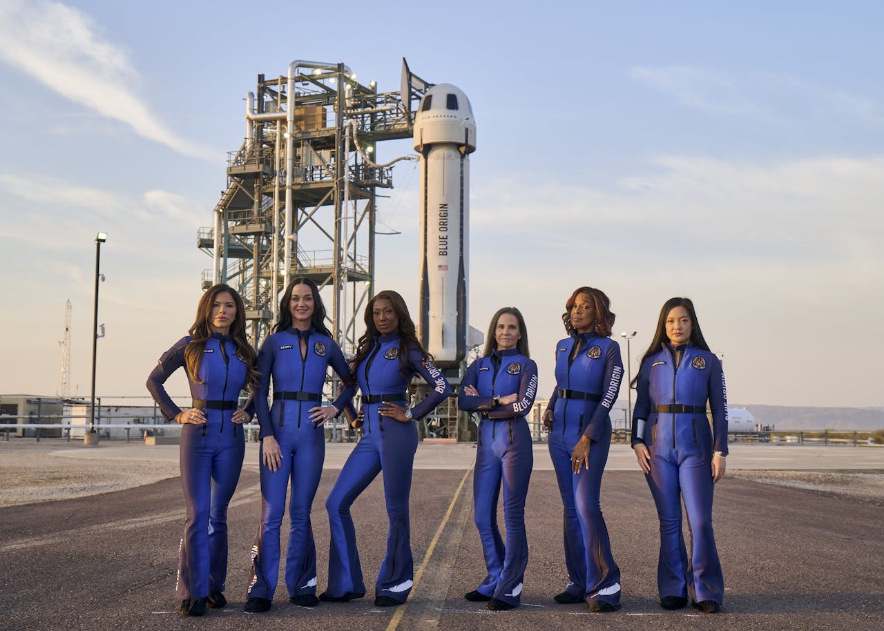 De crew van de elfde bemenste ruimtevlucht van de New Shepard-raket poseert voor de lancering. Zes vrouwen in een blauw pak op asfalt met op de achtergrond een raket op een lanceerplatform.